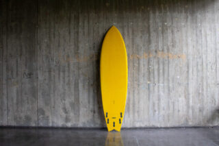 Ein gelbes Surfbrett mit schwarzen Finnen lehnt aufrecht an einer grauen Betonwand mit schwachen Graffiti.