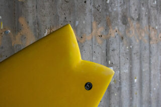 Nahaufnahme des gelben Endes eines fischschwanzförmigen Surfbretts vor einer strukturierten, grauen Betonwand.