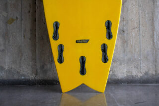 Nahaufnahme der Unterseite eines gelben Surfbretts mit fünf schwarzen Finnenkästen, das an einer rauen Betonwand lehnt.