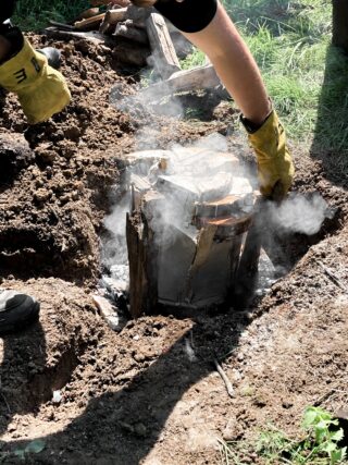 Eine Person mit gelben Handschuhen hebt an einem sonnigen Tag im Freien einen Metallbehälter aus einer Erdgrube, aus der Rauch aufsteigt.