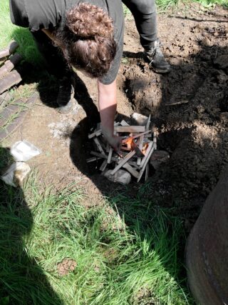 Eine Person hockt im Gras und kümmert sich um ein kleines Feuer, das sie mit Anzündholz und Stöcken in einer flachen Erdgrube gemacht hat.