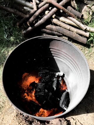 Ein großer Metallbehälter mit brennender Holzkohle darin, im Freien auf Erde platziert, im Hintergrund sind Holzscheite gestapelt.