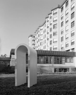 Schwarzweißfoto eines modernen mehrstöckigen Gebäudes mit vielen Fenstern, mit einer minimalistischen Bogenskulptur auf der Rasenfläche im Vordergrund.