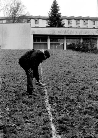 Eine Person markiert eine gerade weiße Linie auf einem grasbewachsenen, laubbedeckten Hang vor einem modernen Gebäude.