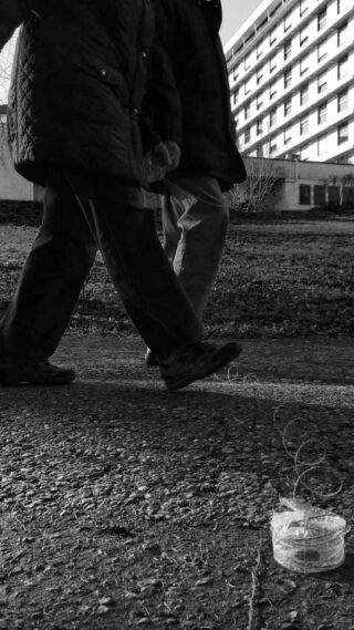 Zwei Personen gehen auf einem gepflasterten Weg an einem Gebäude vorbei. Auf dem Boden steht ein kleiner Behälter, hinter dem sich eine Schnur zieht.