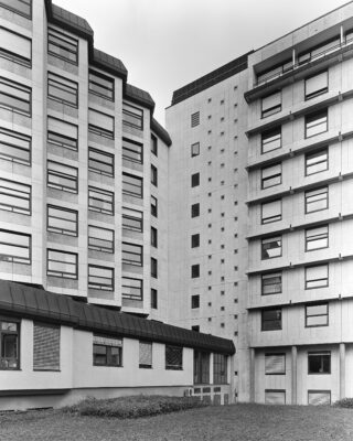 Schwarzweißfoto von zwei modernen, mehrstöckigen Gebäuden mit zahlreichen Fenstern, Flachdächern und einer kleinen Rasenfläche im Vordergrund.