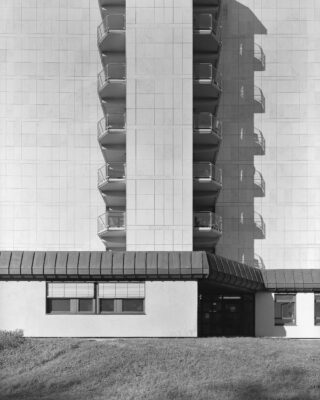 Schwarzweißfoto einer modernen Gebäudefassade mit mehreren Balkonen und einem zentralen Eingang, von vorne betrachtet mit Schattenwurf an der Wand.