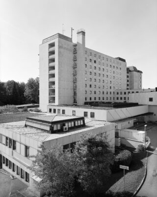 Ein mehrstöckiges Krankenhausgebäude mit einem kleineren Anbau davor, umgeben von Bäumen, fotografiert in Schwarzweiß.