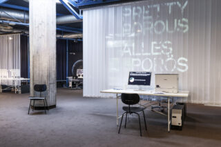 Moderne Büroeinrichtung mit Schreibtisch, Computern und Stühlen in einem Raum im Industriestil mit Sichtbeton und einem großen durchscheinenden Bildschirm, auf dem projizierter Text angezeigt wird.