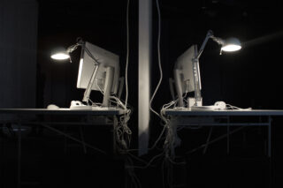 Zwei Computerarbeitsplätze mit Monitoren, Lampen und Kabeln stehen Rücken an Rücken, getrennt durch eine Trennwand, in einem schwach beleuchteten Raum.