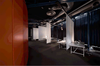 Moderne Büroräume mit dunklem Boden, weißen Schreibtischen, Metallrohren an der Decke, transparenten weißen Raumteilern, dunklen Vorhängen und einer orangefarbenen Wand auf der linken Seite.