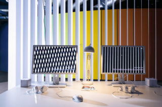 Zwei Computermonitore mit abstrakten Schwarz-Weiß-Mustern stehen auf einem weißen Schreibtisch mit Kopfhörern, Lautsprechern, einer Lampe und einer Maus; im Hintergrund ist eine bunte Wand zu sehen.