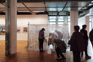 Menschen beobachten und interagieren mit einer Installation aus silbernem Lamettavorhang in einer Kunstgalerie mit gerahmten Kunstwerken an den Wänden.