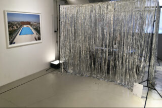 In einem Ausstellungsraum mit Betonboden hängt an einer Galeriewand neben einem Trennvorhang mit metallisch silbernen Fransen.