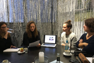 Vier Frauen sitzen mit Getränken und Zeitungen um einen Tisch und unterhalten sich. Vor ihnen auf dem Tisch steht ein geöffneter Laptop mit einem Bild.