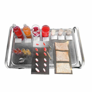 Auf einem Metalltablett befinden sich Flaschen, Gläser mit farbigen Flüssigkeiten, beschriftete Karten, ein Löffel, Tropfen farbigen Gels auf schwarzen Blättern und drei Stücke strukturierter, beiger essbarer Blätter.