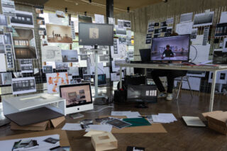 Ein architektonischer Arbeitsbereich mit Computern, maßstabsgetreuen Modellen, Papieren und Designbildern, die die Wände und den Boden bedecken; auf einem der Computerbildschirme ist eine Person zu sehen.