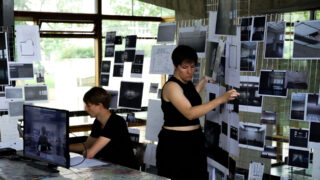 Zwei Personen arbeiten in einem Studio mit Architekturplänen und Fotografien, die auf Tafeln und Wänden angezeigt werden. Einer sitzt an einem Schreibtisch mit einem Computer, der andere ordnet Bilder auf einem Anzeigeraster an.