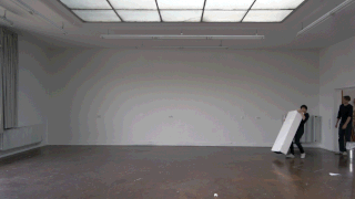 Zwei Personen befinden sich in einem leeren Raum mit weißen Wänden und einem großen Oberlicht; eine Person trägt einen großen weißen rechteckigen Gegenstand.