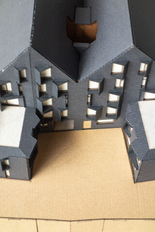 Ein Architekturmodell aus Karton eines Gebäudes mit unregelmäßig angeordneten Fenstern und zwei kleineren Baukörpern, die auf einen offenen Innenhof blicken.
