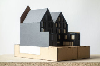 Auf einem einfachen Sockel steht ein maßstabsgetreues Architekturmodell eines modernen mehrstöckigen Gebäudes mit dunklen Außenwänden.