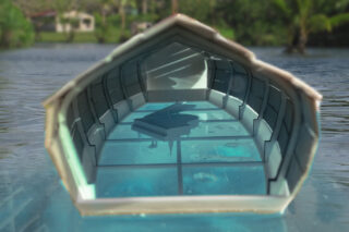 Eine kleine, teilweise geschlossene Glaskonstruktion schwimmt auf dem Wasser. Im Inneren steht auf einem transparenten Boden ein Flügel. Im unscharfen Hintergrund sind Bäume und Gebäude zu sehen.