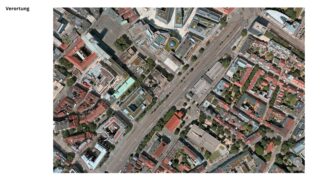 Luftaufnahme eines Stadtgebiets mit Straßen, Gebäuden und Grünflächen, wobei eine große Straße diagonal durch die Mitte verläuft.