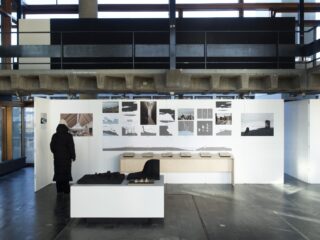 Eine Person betrachtet Architekturzeichnungen und Modelle, die auf weißen Wänden und Tischen in einem modernen Ausstellungsraum mit Beton- und Glaselementen ausgestellt sind.