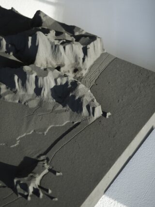 Ein graues topografisches Modell weist strukturierte Hügel und Bergrücken auf, die von feinen Linien durchzogen sind, die Straßen oder Wege andeuten, und steht auf einer rechteckigen Basis mit einer schattigen Ecke.