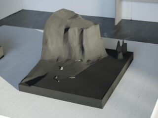 Ein dunkelgraues Architekturmodell zeigt einen großen, felsigen Hügel mit kleinen verstreuten Strukturen auf einer rechteckigen Basis, die auf einem weißen Tisch ausgestellt ist.