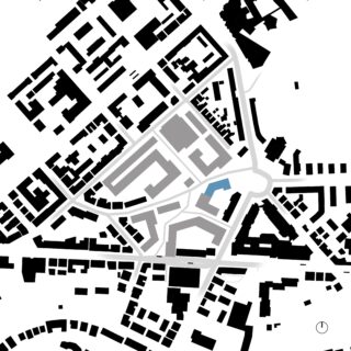 Schwarz-weiße schematische Karte eines Stadtgebiets mit Straßen, Gebäuden, einem Kreisverkehr und einem blau hervorgehobenen Gebäude in der Mitte rechts. Unten rechts befindet sich ein kleiner Kompass.