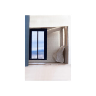 Ein kleiner Raum mit einem blau gerahmten Fenster, offenen Fensterläden und einem Stück weißem Stoff, das an der rechten Wand hängt.