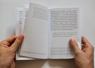 Zwei Hände halten ein Buch und blättern es durch, das auf einer Doppelseite mit deutschem Text aufgeschlagen ist. Der Blick fällt auf Seiten mit dicht gedrucktem Inhalt.
