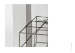 Eine Gitterstruktur aus Metalldraht befindet sich neben einer weißen vertikalen rechteckigen Säule vor einem schlichten weißen Hintergrund.