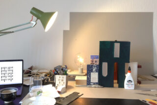 Ein überfüllter Schreibtisch mit einer leuchtenden grünen Schreibtischlampe, einem Laptop, einem Glas Wasser, Klebstoff, Architekturmodellen, einer Glühbirne und verstreutem Briefpapier.