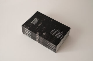 Ein Stapel schwarzer Softcover-Zeitschriften mit dem Titel „Samenin“ und weißem Text auf dem Cover, ordentlich auf einer hellen Oberfläche angeordnet.