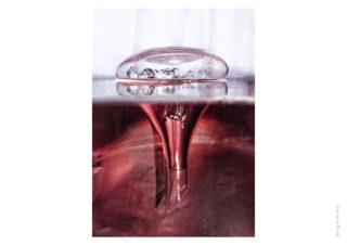 Eine klare Blase ist teilweise in eine rot gefärbte Flüssigkeit eingetaucht, mit einer Spiegelung und einer Flüssigkeitssäule, die sich unter der Oberfläche nach unten erstreckt.