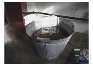 Ein großer, mit Wasser gefüllter Metalleimer, in dem sich ein Ziegelstein, ein Glasgefäß und ein weißes Tuch befinden; daneben stehen ein grüner Schlauch und ein Saftkarton.