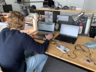 Eine Person sitzt an einem Schreibtisch und arbeitet an zwei Laptops. Vor ihr auf dem Tisch liegen elektronische Komponenten und Kabel.