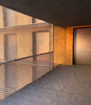 Nahaufnahme eines Fliegengitters, das ein offenes Fenster oder einen Balkonbereich abdeckt, wobei das Sonnenlicht warme Töne auf strukturierte Beton- und Ziegeloberflächen wirft.