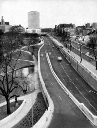 Schwarzweißfoto einer geteilten Stadtstraße mit Autos, neben einem Kanal, Bäumen und Gebäuden aus der Mitte des Jahrhunderts im Hintergrund.