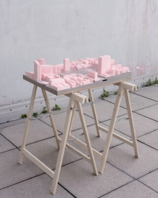 Auf einem hölzernen Sägebocktisch auf einer gefliesten Außenfläche in der Nähe einer weißen Wand wird ein maßstabsgetreues Modell einer Stadt mit rosafarbenen Gebäuden ausgestellt.