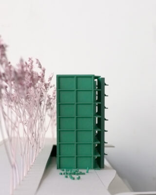 Ein grünes Architekturmodell eines rechteckigen Gebäudes steht vertikal neben einer Reihe rosafarbener Miniaturbäume auf einem weißen Sockel.