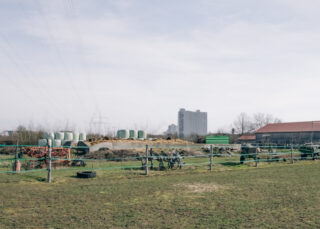Ein eingezäuntes Grasfeld mit landwirtschaftlichen Geräten, in weiße Plastikfolie eingewickelten Heuballen und einem entfernten Wohnhaus unter einem dunstigen Himmel.