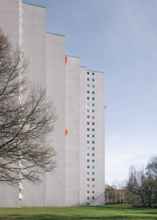 Ein hohes, weißes Wohngebäude mit stufenförmigem Grundriss und orangefarbenen Akzenten auf einigen Balkonen steht neben blattlosen Bäumen und einer Rasenfläche unter klarem Himmel.
