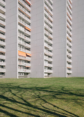 Hohes, graues Wohnhaus mit mehreren Balkonen und orangefarbenen Markisen, hinter einer grünen Rasenfläche mit Baumschatten.