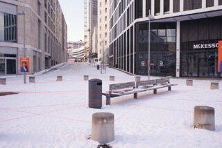 Ein schneebedeckter Stadtplatz mit Bänken und Pollern, verbunden durch rote Schnüre; im Hintergrund sind moderne Gebäude und ein Kran zu sehen.
