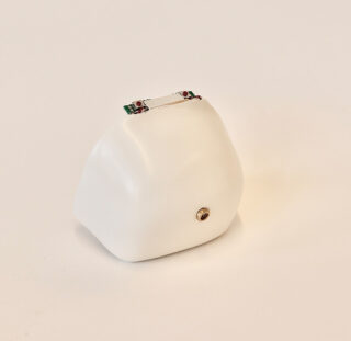 Ein kleines, weißes, unregelmäßig geformtes Objekt mit einer metallischen Komponente oben und einem runden Metallteil auf einer Seite, platziert auf einem schlichten weißen Hintergrund.