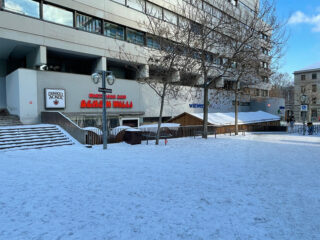 Schnee bedeckt den Boden vor einem modernen Gebäude mit Schildern für Duckre Packe und Brauhaus, einigen kahlen Bäumen und Fahrrädern, die in der Nähe der Straße geparkt sind.