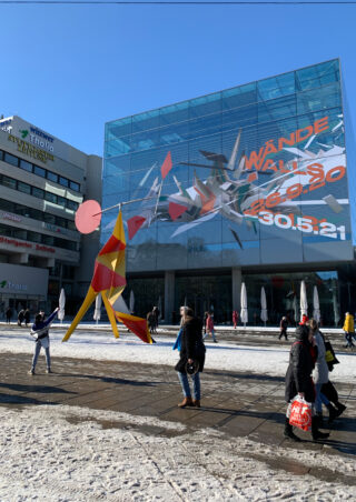 Menschen gehen auf einem verschneiten Platz vor einem Glasgebäude mit farbenfroher abstrakter Kunst und großem Text an der Fassade spazieren; im Vordergrund steht eine moderne Skulptur.